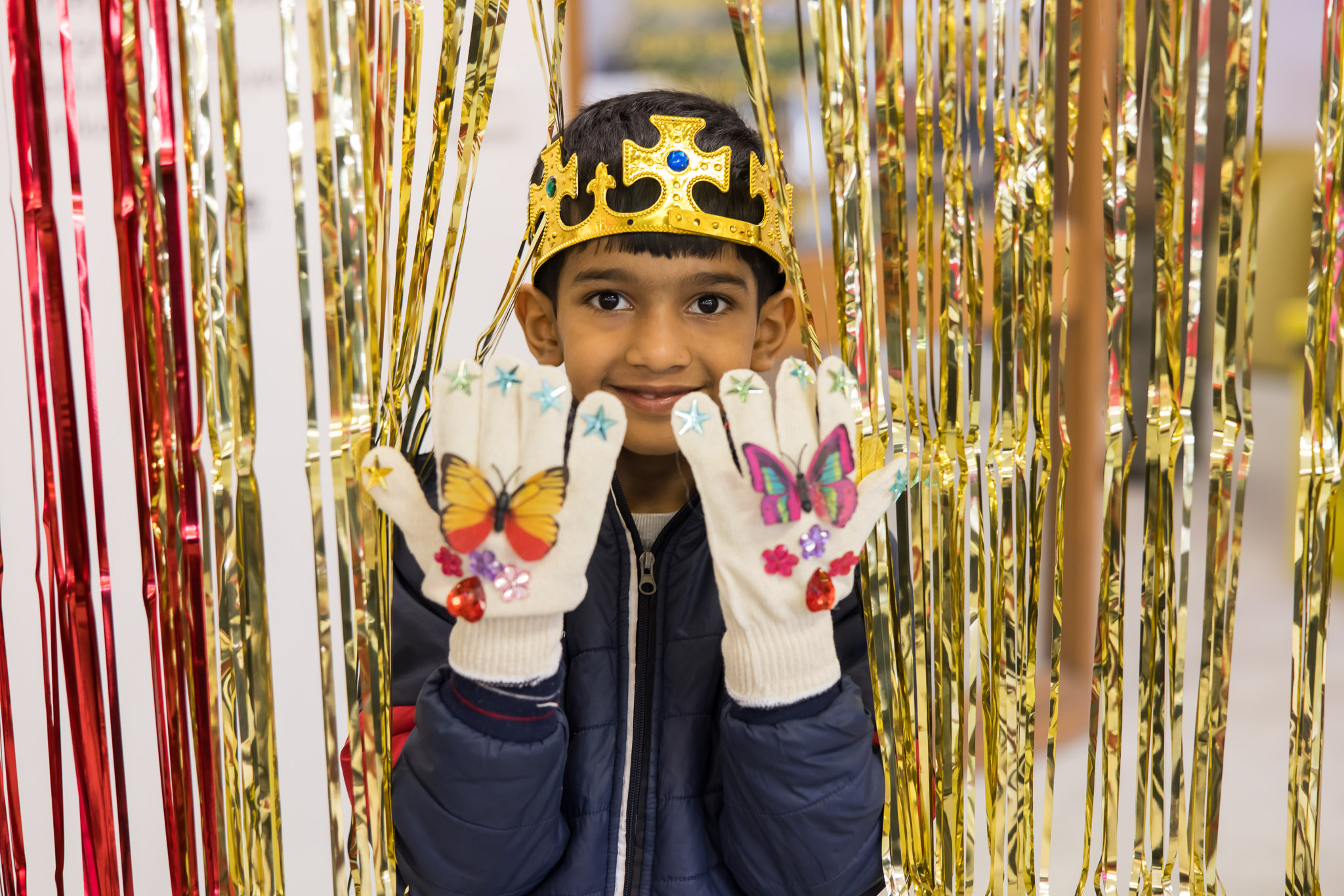 A child wearing a crown shows their decorated gloves while standing in a red and gold tinsel curtain