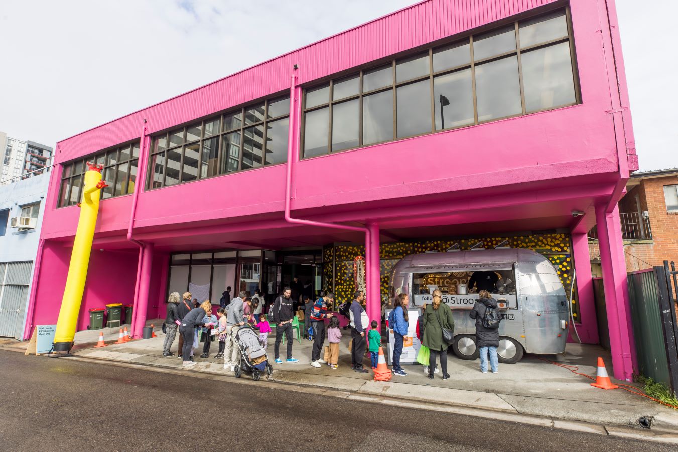 People gather outside a large, bright pink building. There is a silver food truck and a tubular yellow air dancer man