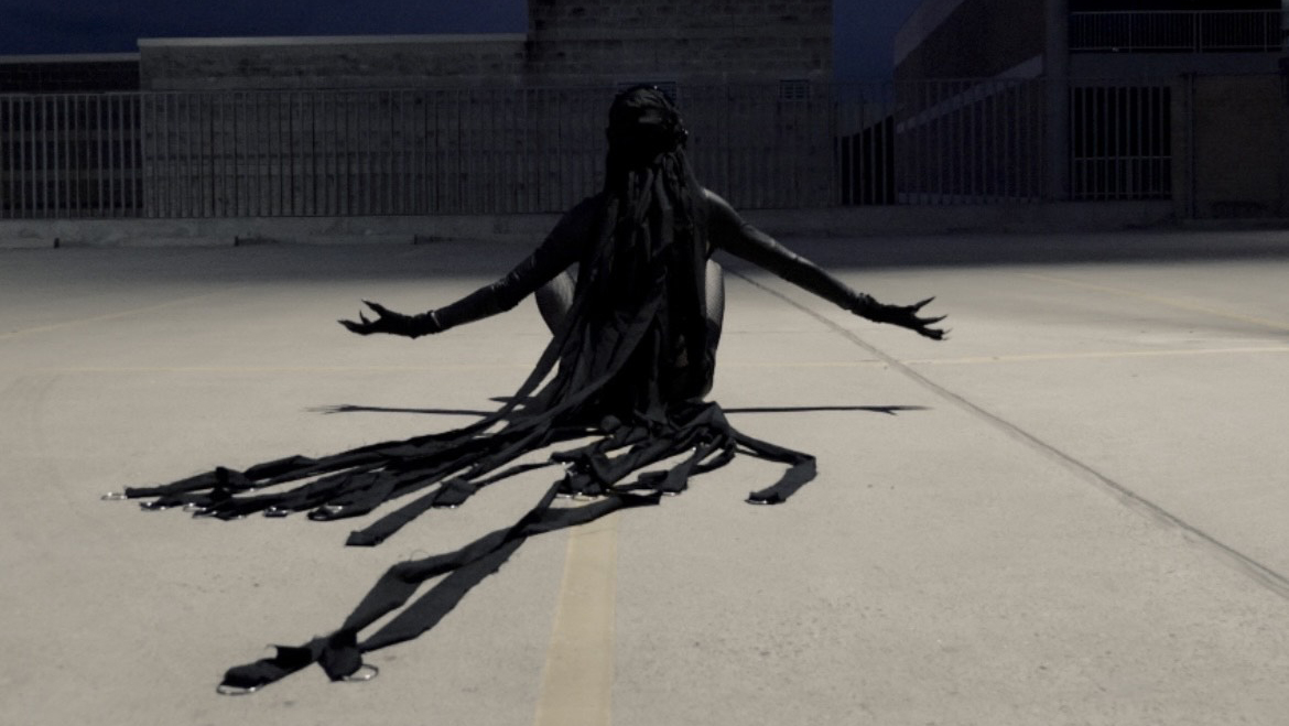 A black costumed figure sits cross legged facing away from camera