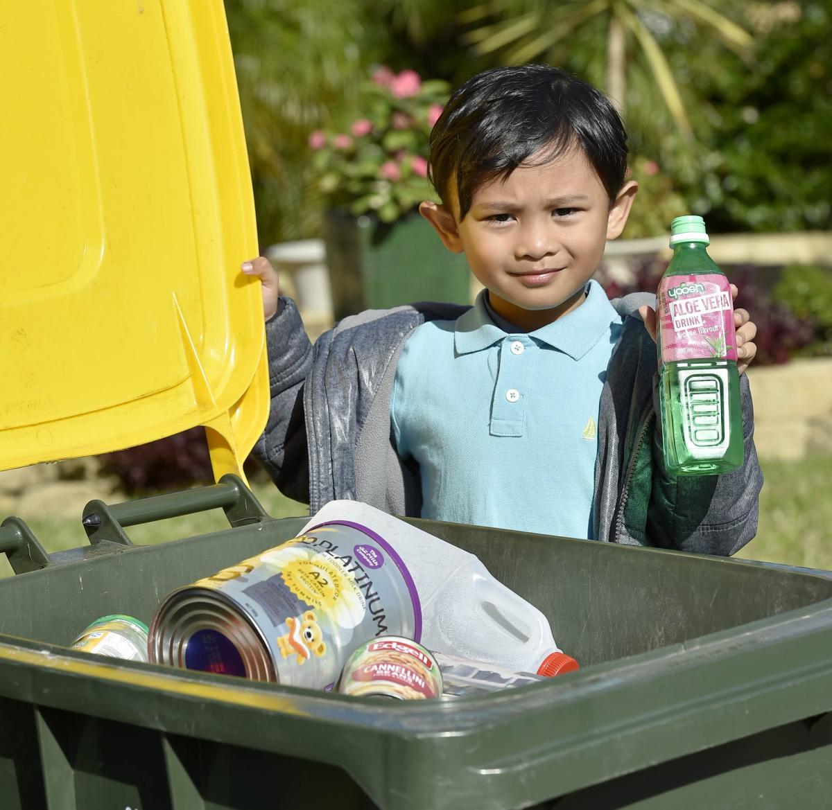 Little boy holding aloe vera water bottle next to yellow recycling bin