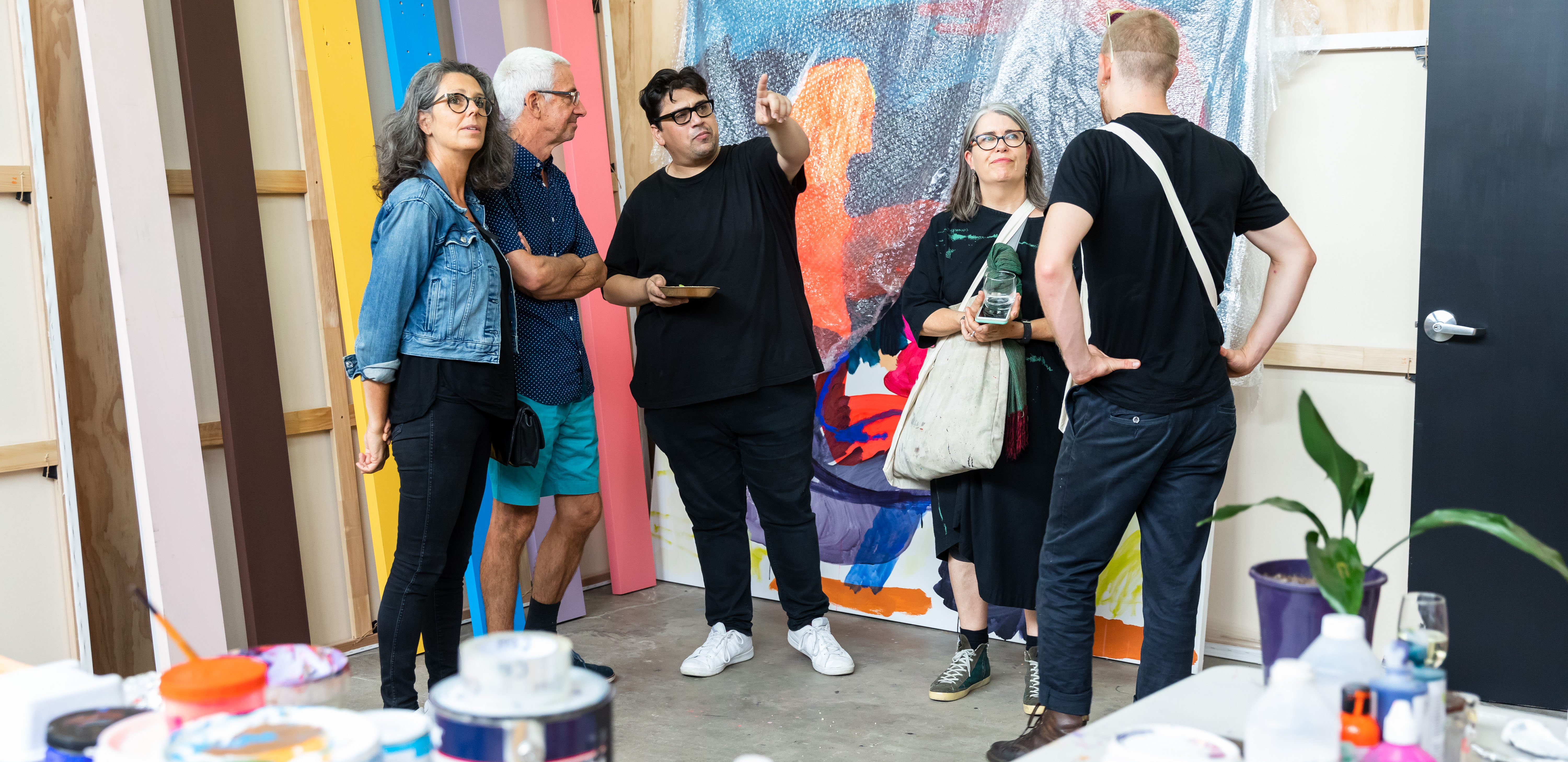 A group of people gathered talking in the corner of a studio filled with brightly coloured paintings