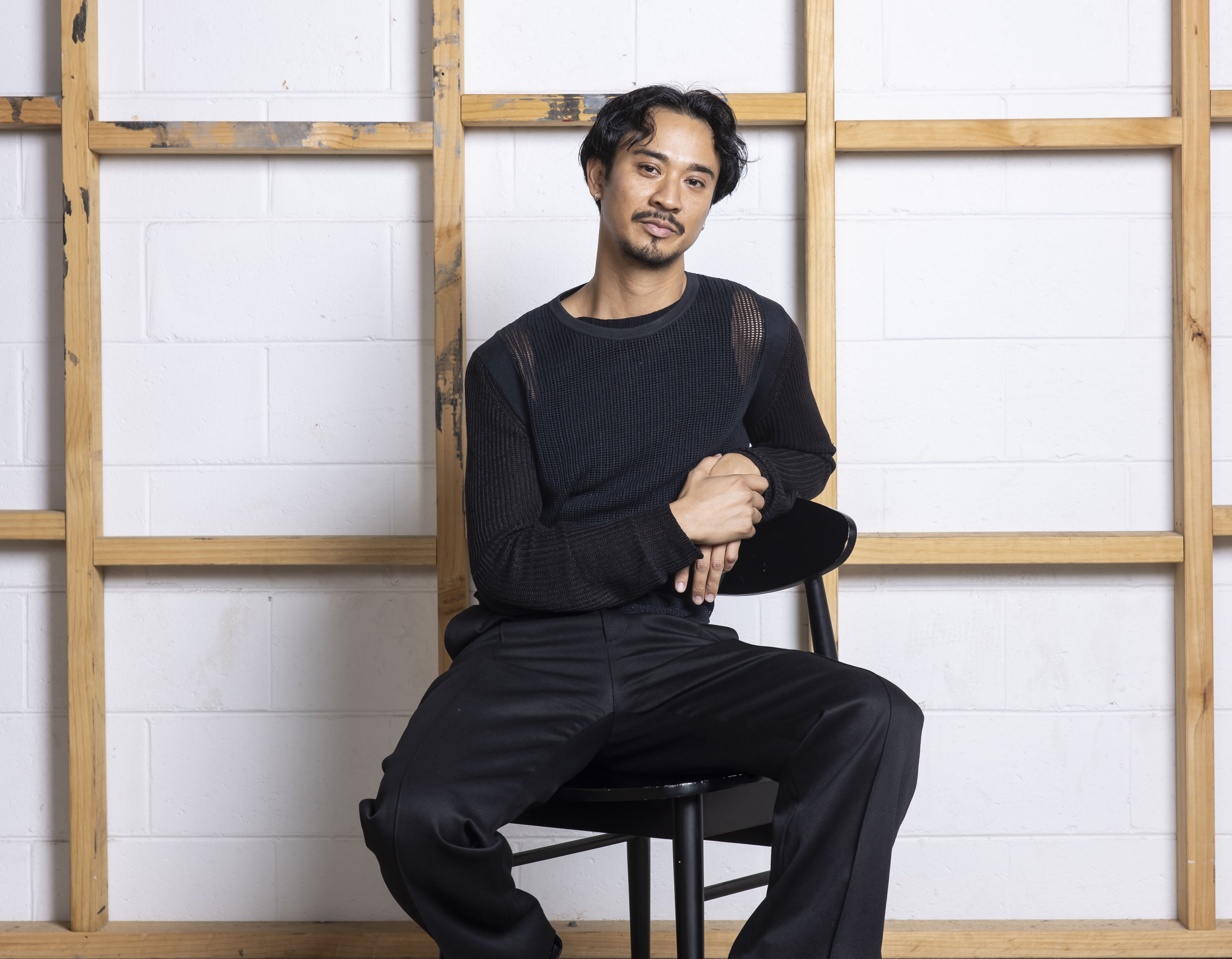 A man with black hair in dark clothing sits on a black chair against a wooden stud wall