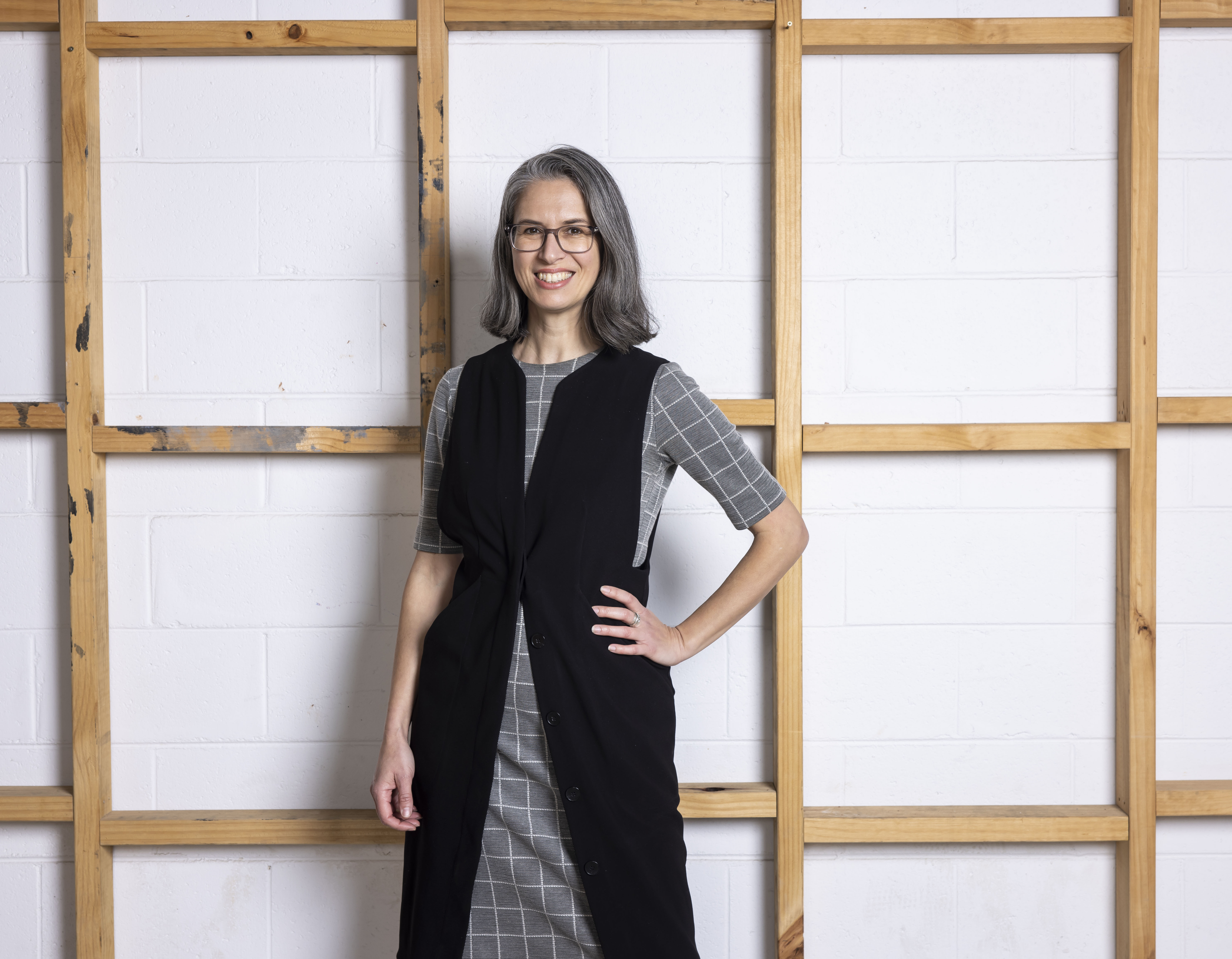 A woman with a grey bob wearing a grey dress with a long black sleeveless jacket standing against a wooden stud wall