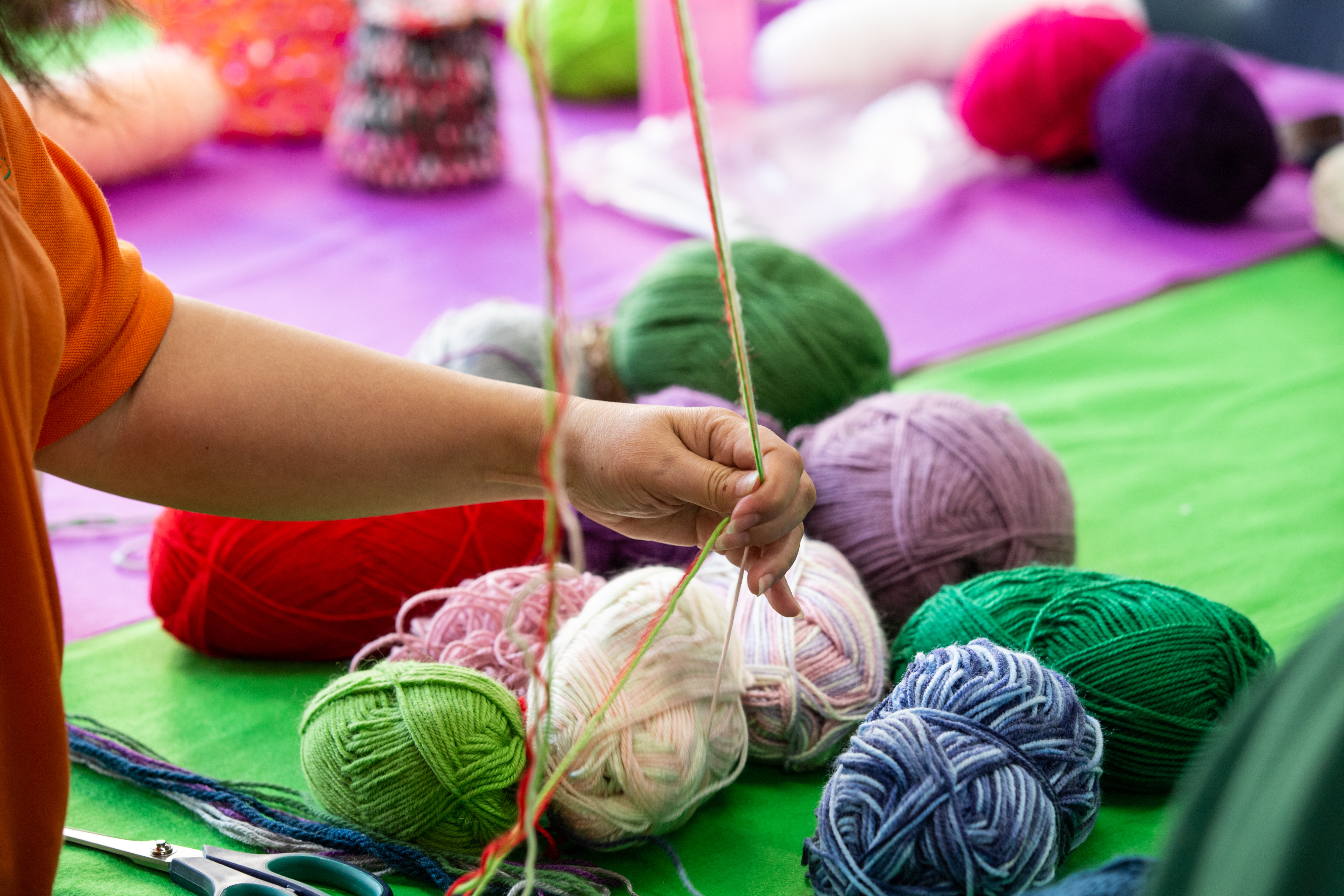 A person stretches wool with their hands. Their are many balls of colourful wool in a pile on a table in the background.